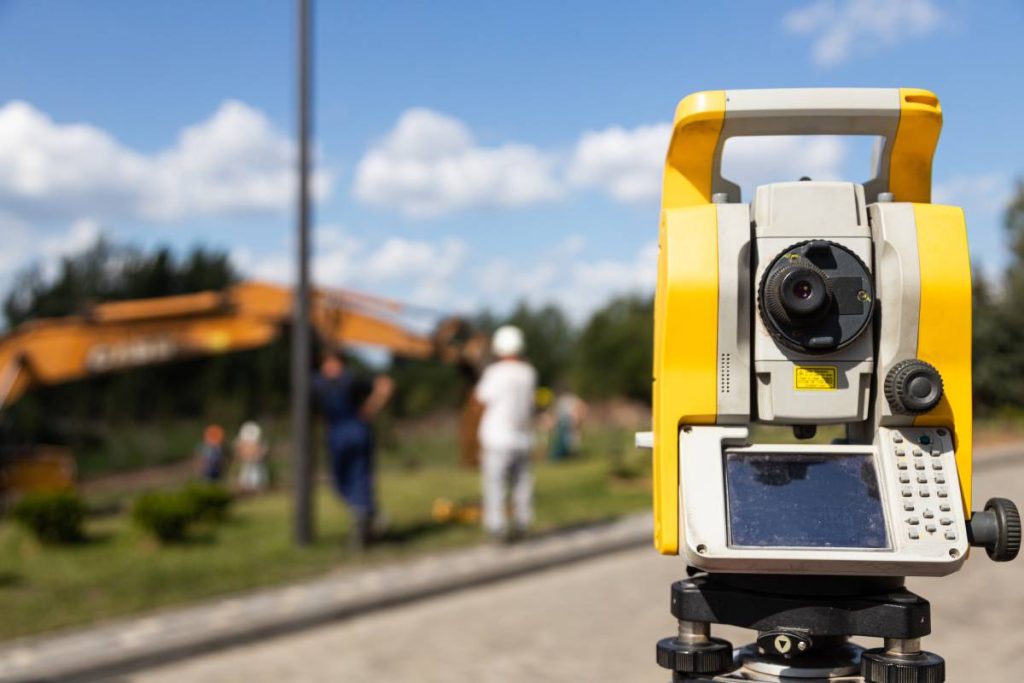 Close-up of yellow and orange tacheometer standing on street against work on construction site. Photo with depth of field