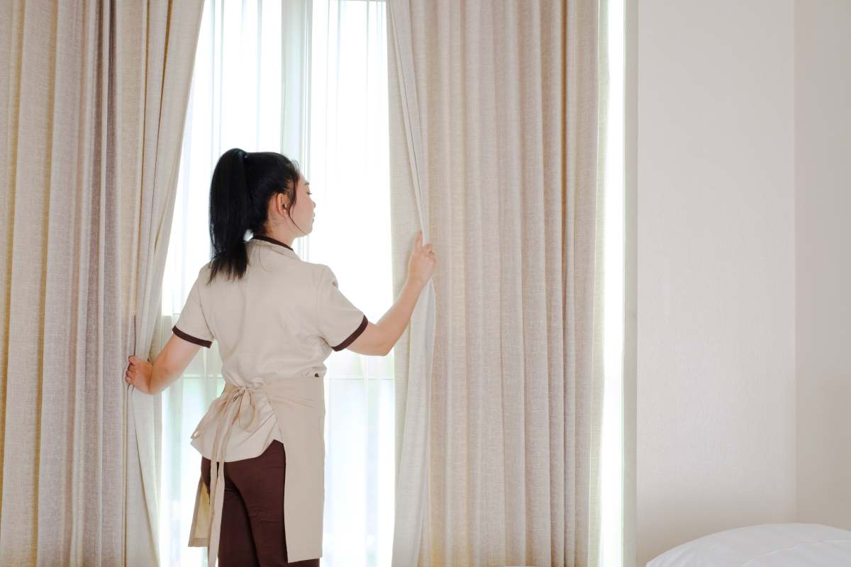 Young maid opening curtains in hotel room