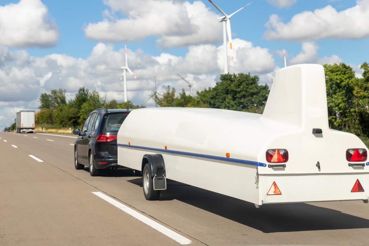 Big white disassembled glider plane in cover box on trailer towed by van or suv on highweay road sunny day against blue sky. Sailplane equipment transport and storage. Extreme leisure hobby activity.