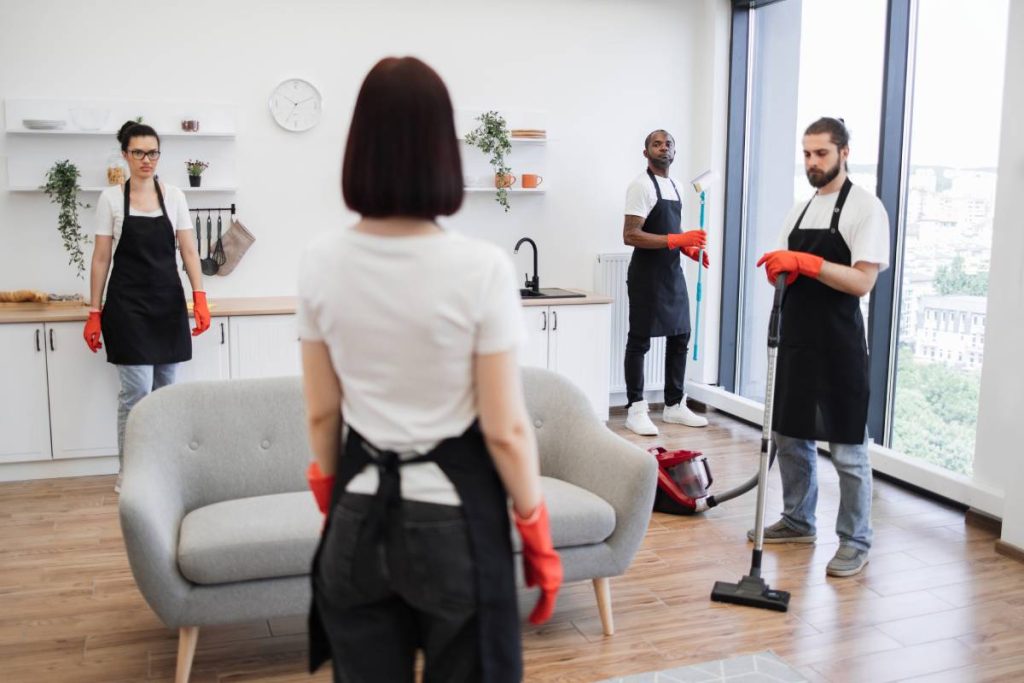 Back view of female boss of cleaning company, telling work schedule to cleaners of studios spacious kitchen. Group of janitors in uniform cleaning office with cleaning equipment.