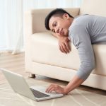 Lazy young Vietnamese man lying on sofa and watching something on laptop