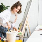 Young inspired girl dunks a paintbrush into the paint in light studio