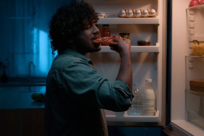 Home kitchen at night dark room hungry Hispanic man Indian male guy want eat rubbing belly open fridge searching food meal in refrigerator insomnia addiction unhealthy habit eating slice of pizza. High quality photo